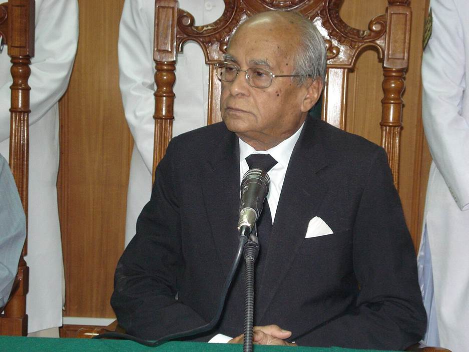 A person in a suit sitting at a microphone  Description automatically generated